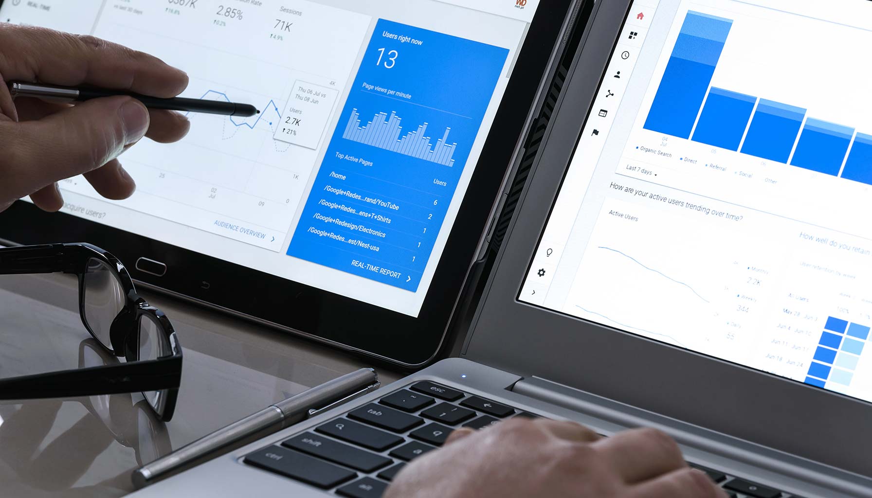 Two laptops display analytics dashboards with charts and graphs. A hand holds a pen pointing at one screen. Glasses and a pen are on the table.